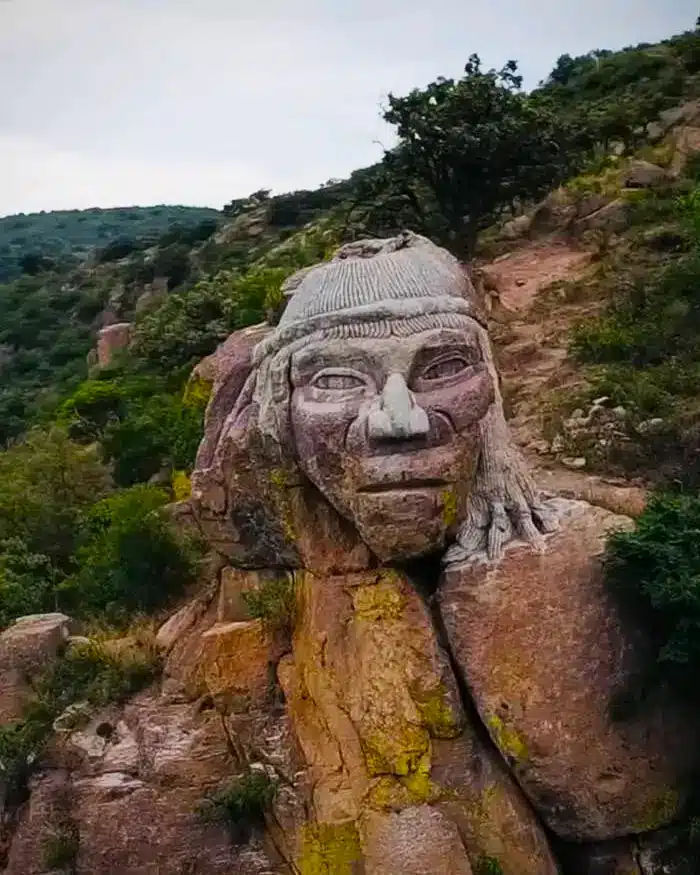 La escena muestra una cabeza monumental de piedra ubicada en una zona natural con cerros, árboles y vegetación alrededor.