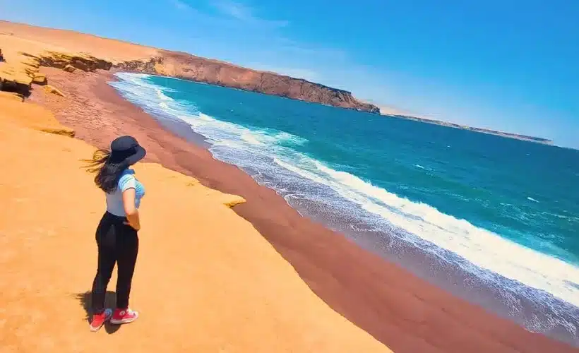 A panoramic view of Playa Roja in Paracas, Peru, showing its striking red sand caused by igneous rock erosion, contrasting with deep blue ocean waves and desert cliffs.