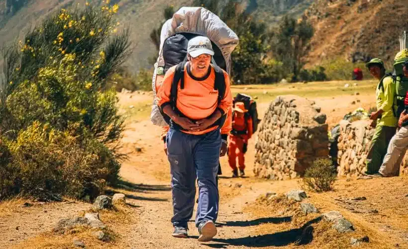 Porters carry camping equipment and supplies along the Inca Trail and Salkantay Trek near Cusco, Peru, surrounded by green mountains and cloudy skies.