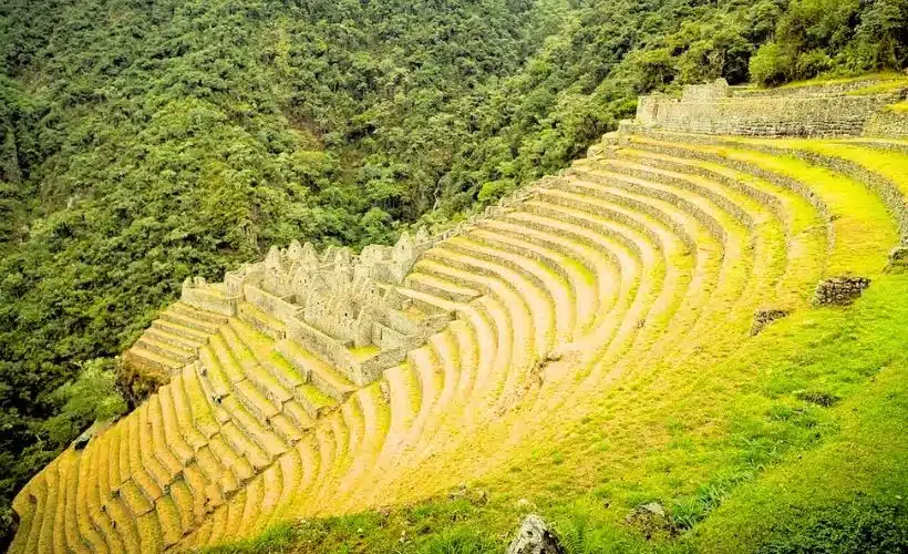 Aerial view of the Wiñay Wayna archaeological site along the Inca Trail near Machu Picchu, showing Inca terraces, stone ruins, and lush green Andean mountains under the sun.