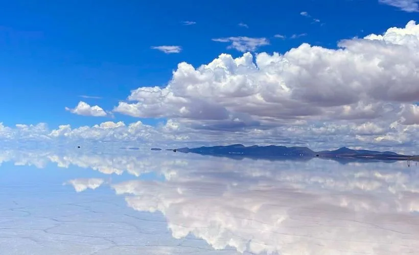 Panoramic view of the Uyuni Salt Flats under a cloudy sky, showing the mirror effect during the rainy season in Bolivia, reflecting tourists and distant mountains.