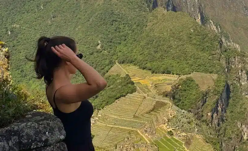 A traveler standing on the summit of Huchuy Picchu, overlooking Machu Picchu citadel and the Urubamba Valley below, under bright sunlight and clear blue skies.