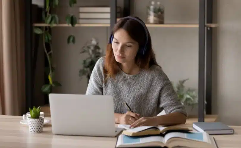 Woman at a wooden table uses laptop and phone beside a coffee cup, booking Machu Picchu tickets and transport from Cusco to Aguas Calientes with agency support.