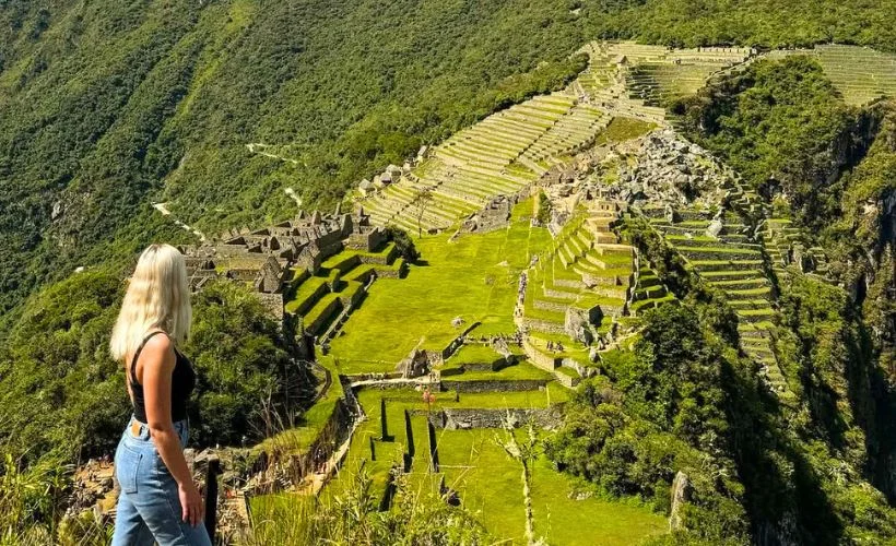 Woman in trekking clothes walks a well-marked path on Huchuy Picchu, Machu Picchu site, Cusco. Gentle terrain, scattered shrubs, and bright daylight create a calm family-friendly route.