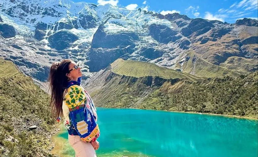 Visitors wearing trekking boots rest beside turquoise Humantay Lake below Salkantay glacier, snowy peaks and rocky shore reflected in calm water on a sunny Cusco morning.