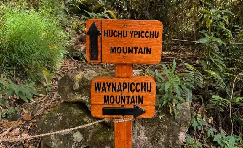 wooden trail sign marking the entrance to the Huchuy Picchu path surrounded by green vegetation and mountains inside the Machu Picchu Sanctuary, Cusco.