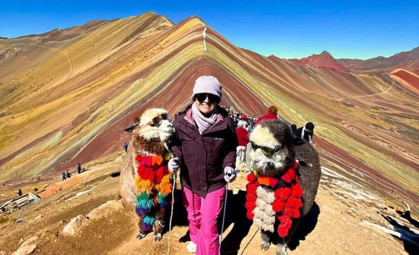Hikers in jackets stand before Vinicunca Rainbow Mountain’s striped slopes at 5,000 m, with llamas nearby, blue sky, scattered clouds, and distant Ausangate in Cusco.
