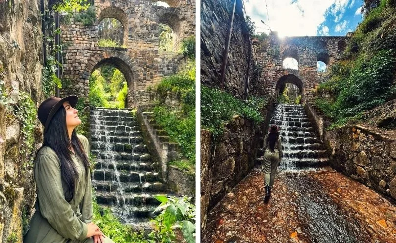 People walk beneath Sapantiana Aqueduct’s stone arches and stairway, framed by greenery and shadows; a photogenic colonial structure near Cusco’s center, perfect for Instagram shots.