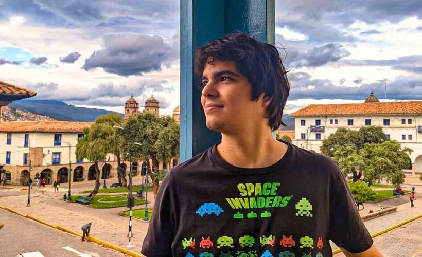 A traveler overlooks Plaza Regocijo’s gardens, colonial arcades, and museum façades in Cusco’s historic center, photographing monuments beneath bright sun and scattered clouds.