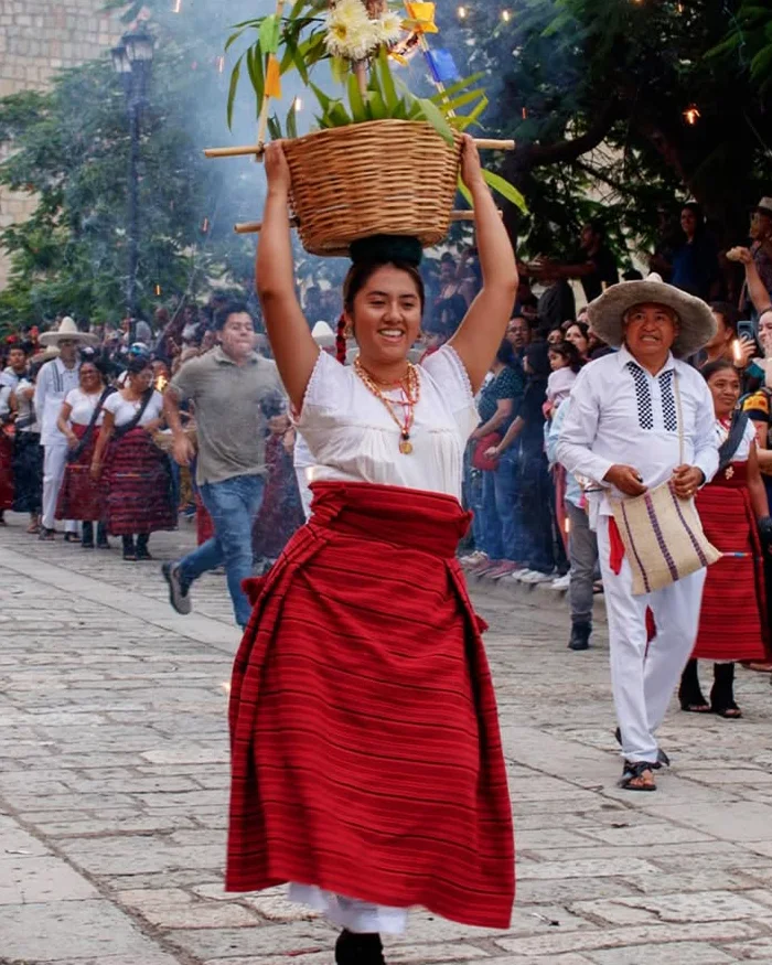 Groups of dancers in vibrant traditional outfits perform synchronized steps during La Guelaguetza festival, surrounded by crowds enjoying music, colors, and regional cultural displays.