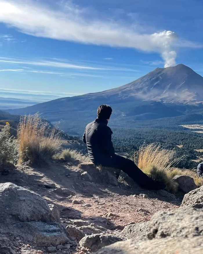 Sentado en la ladera, el viajero, entre pastizales de altura y rocas volcánicas, contempla el Popocatépetl.