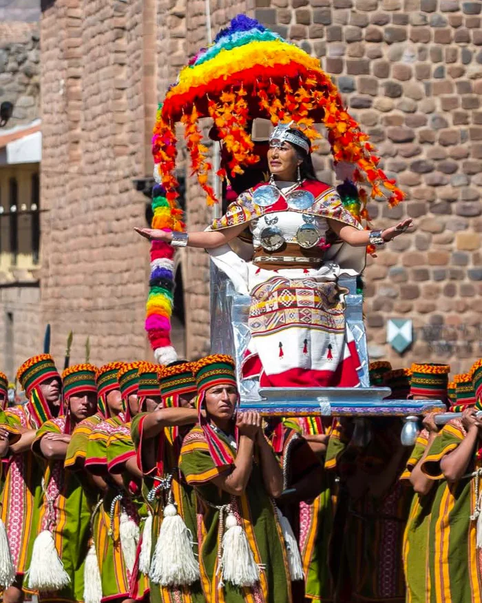 The Coya character walks through Cusco’s main square dressed in ornate traditional Andean clothing with golden details, greeting spectators during the Inti Raymi celebration.