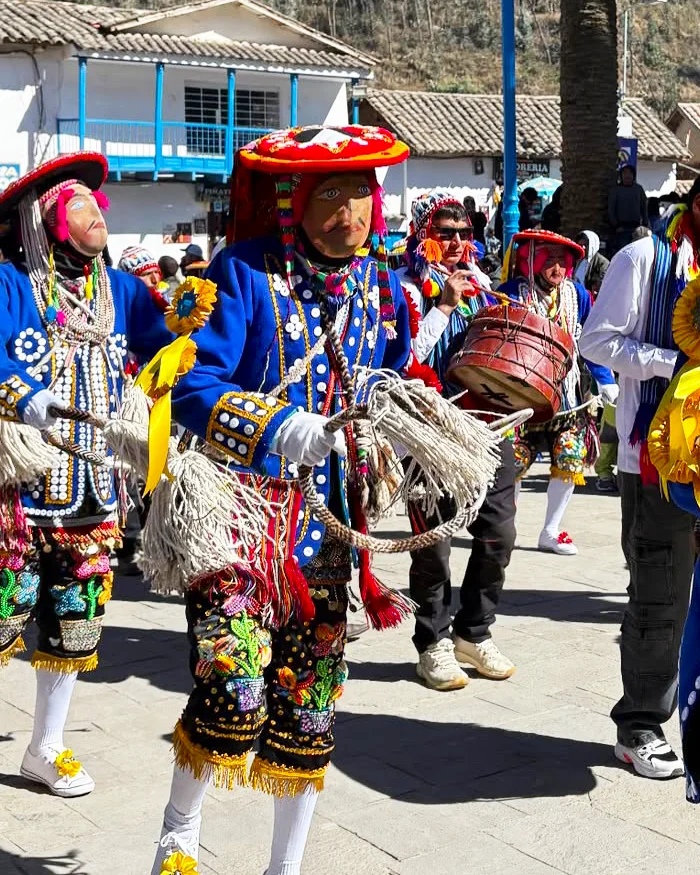 Performers dressed in colorful Andean costumes dance energetically in a public square, showcasing traditional Kachampa choreography with decorative hats, embroidered clothing, and lively movements.