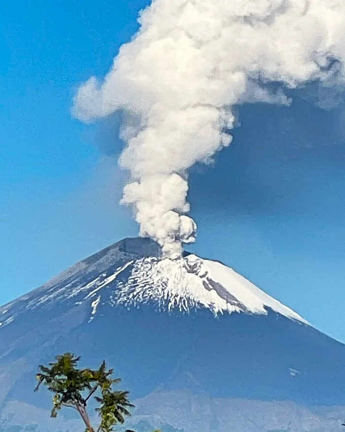 A tall column of white volcanic smoke rises from a crater into a bright blue sky, with surrounding terrain showing green slopes and rocky Andean formations.