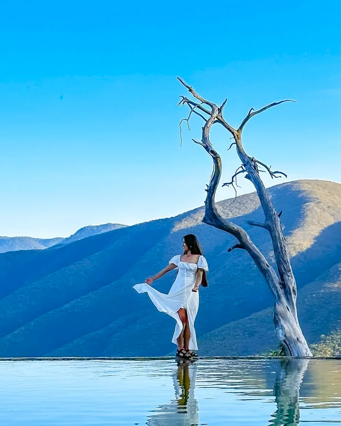 Una viajera vestida con un elegante vestido blanco se encuentra de pie sobre el borde del agua, admirando el paisaje montañoso.