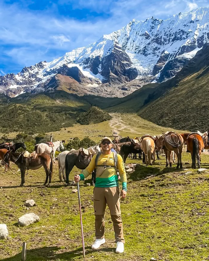 Un viajero posa con bastón de trekking frente a un grupo de caballos, con las imponentes montañas nevadas del Salkantay al fondo.