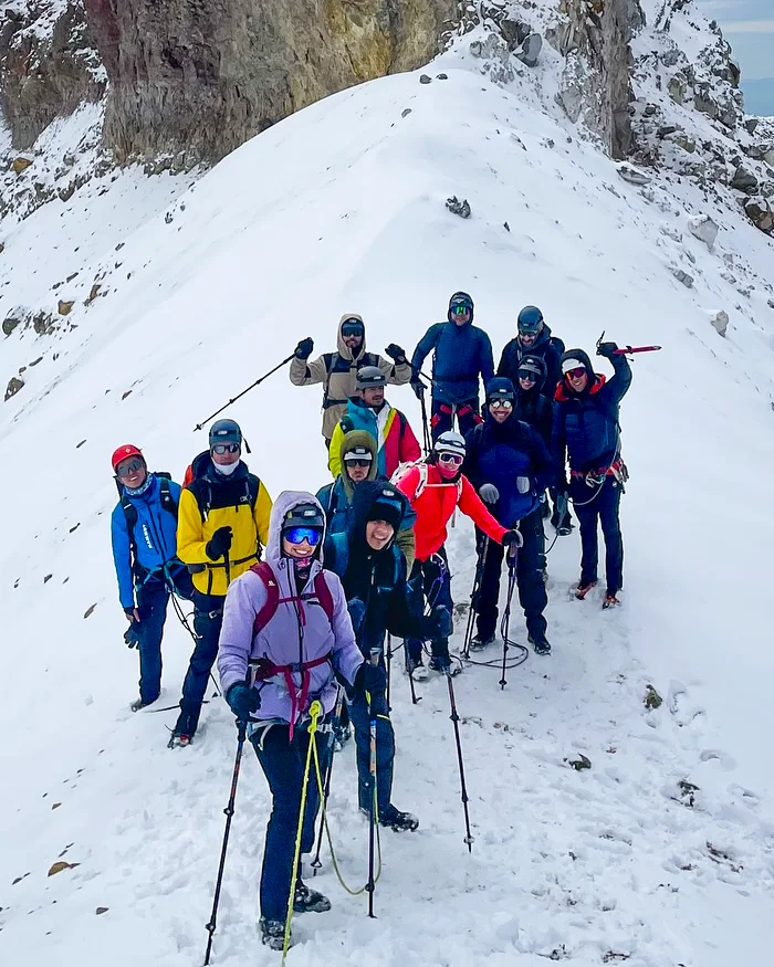 A vertical rock formation stands sharply against snowy Andean terrain, with hikers walking nearby under clear skies that highlight the dramatic high-altitude landscape.