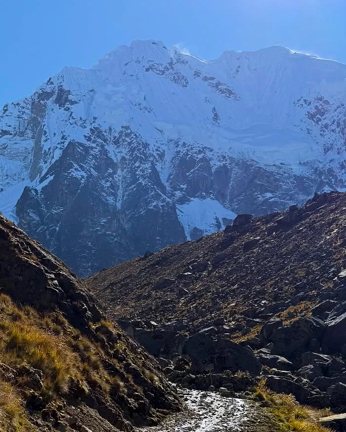 The Salkantay snow-capped peak towers above rolling Andean terrain, with scattered clouds and golden sunlight illuminating the trekking route in Cusco.