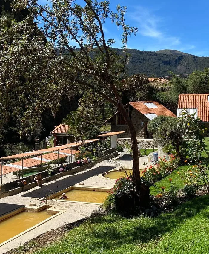 Golden hot spring pools reflect the sunlight while surrounded by mountains, rocks, and clear skies, creating a warm and inviting natural atmosphere.