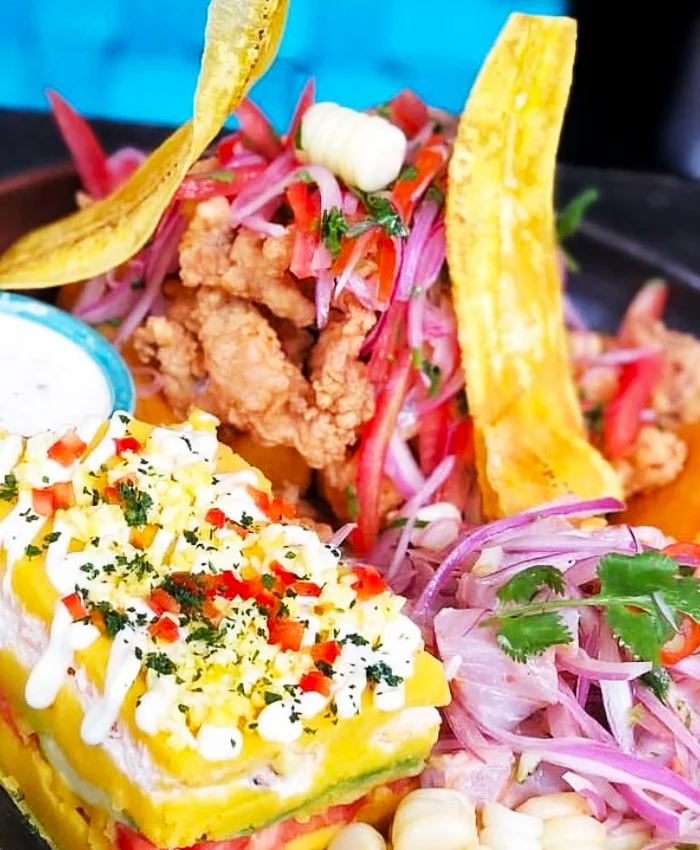 A un costado hay una porción de ceviche, con pescado fresco marinado en jugo de limón, cebolla morada, ají y hojas de cilantro.