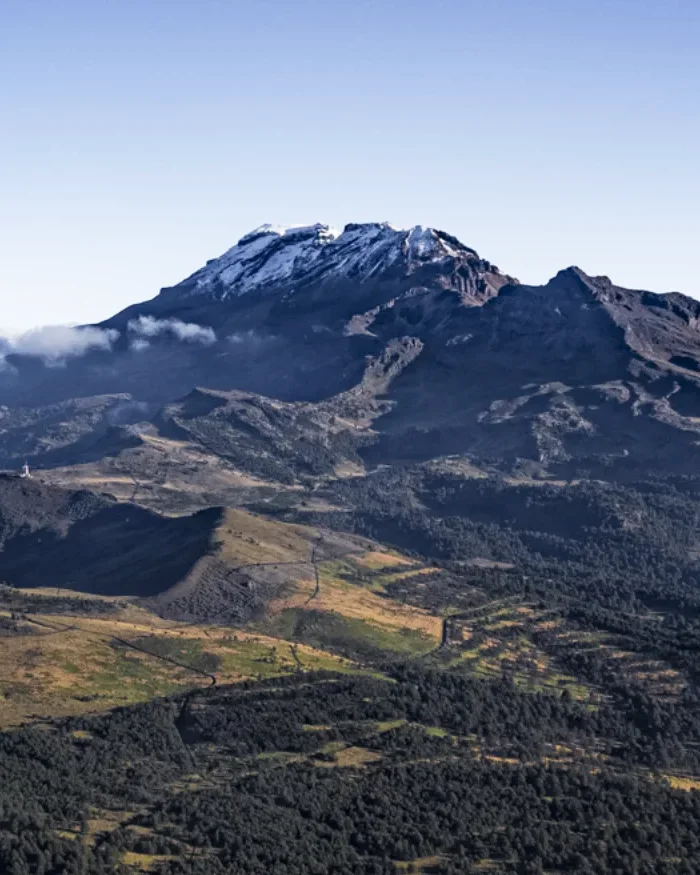 Snow-covered Iztaccíhuatl Mountain stands beneath a clear sky, surrounded by patches of green vegetation and rugged terrain that emphasize its scale and natural beauty.