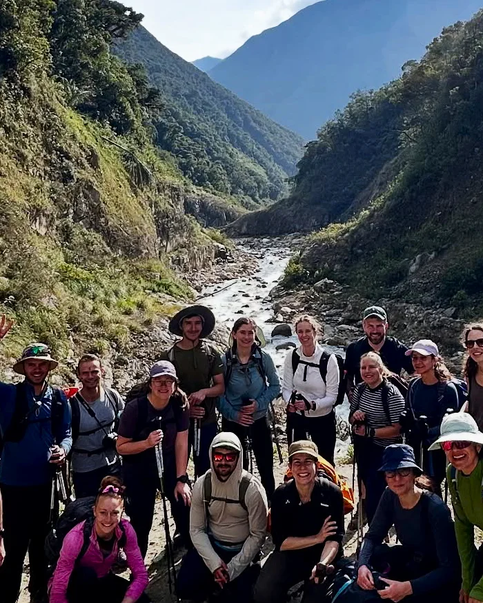 A diverse group of hikers walks along an Andean trail surrounded by mountains and a river, highlighting adventure, nature, and camaraderie in the Cusco region.