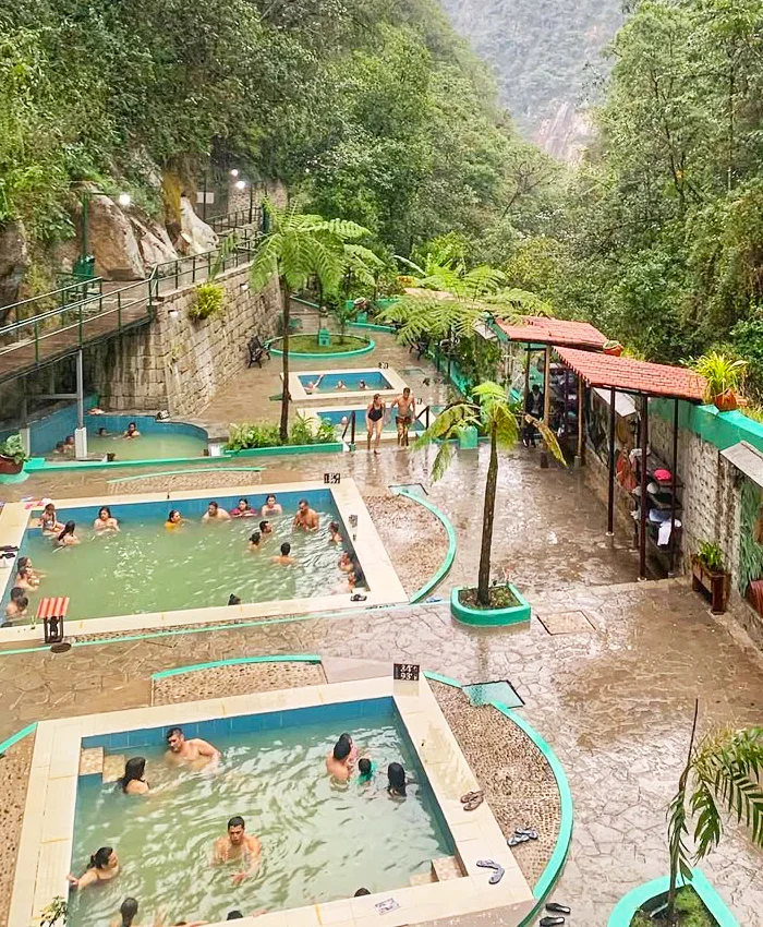 Visitors relax in warm outdoor hot springs bordered by green vegetation, natural rocks, and shaded areas, enjoying the calm atmosphere and soothing water.