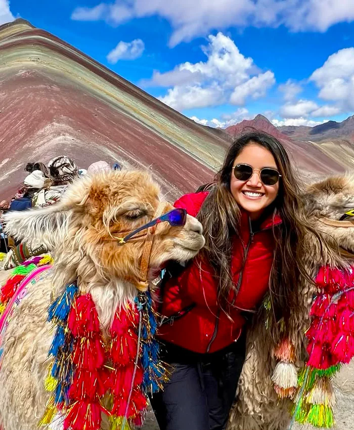 La viajera sonriente posando junto a dos llamas decoradas con coloridos adornos andinos.