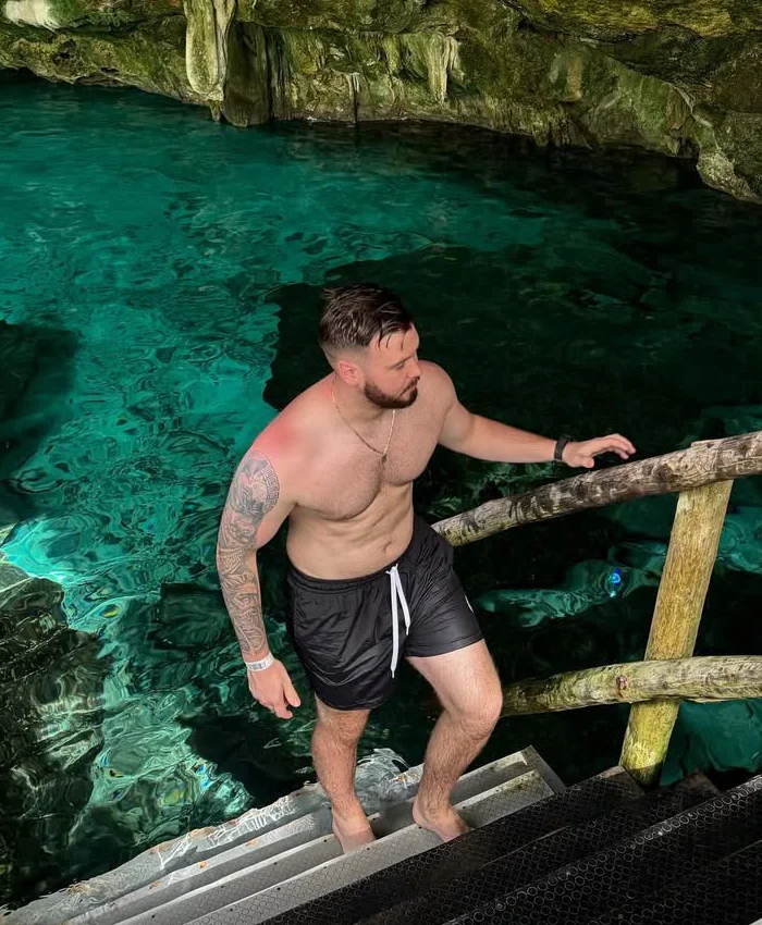 A man sits in a natural pool with clear water, leaning against rock walls, surrounded by soft light and greenery, enjoying a peaceful and contemplative cenote experience.