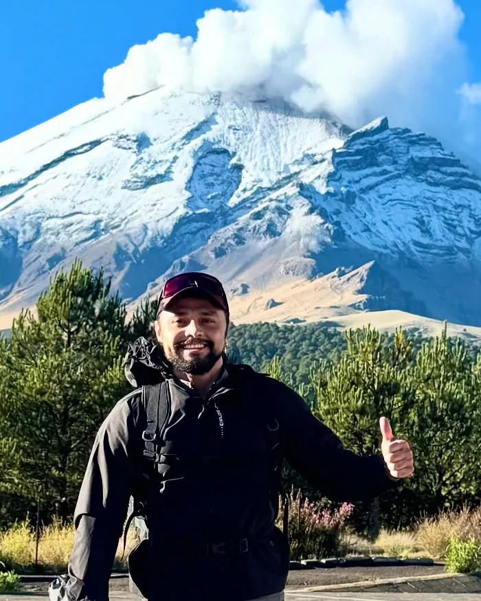 A towering snow-capped volcano emits a white smoke column into the sky, surrounded by green vegetation and contrasting rocky slopes typical of high-altitude Andean regions.