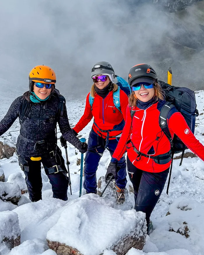 Two women wearing red thermal jackets and safety helmets stand on a snowy Andean trail near Salkantay, with misty mountain scenery typical of high-altitude Cusco landscapes.