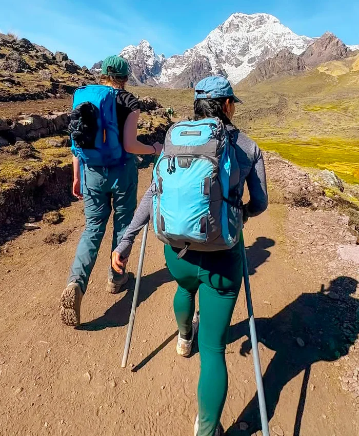 Dos viajeros disfrutan una jornada de trekking en Cusco, caminando entre montañas andinas hacia un nevado impresionante.
