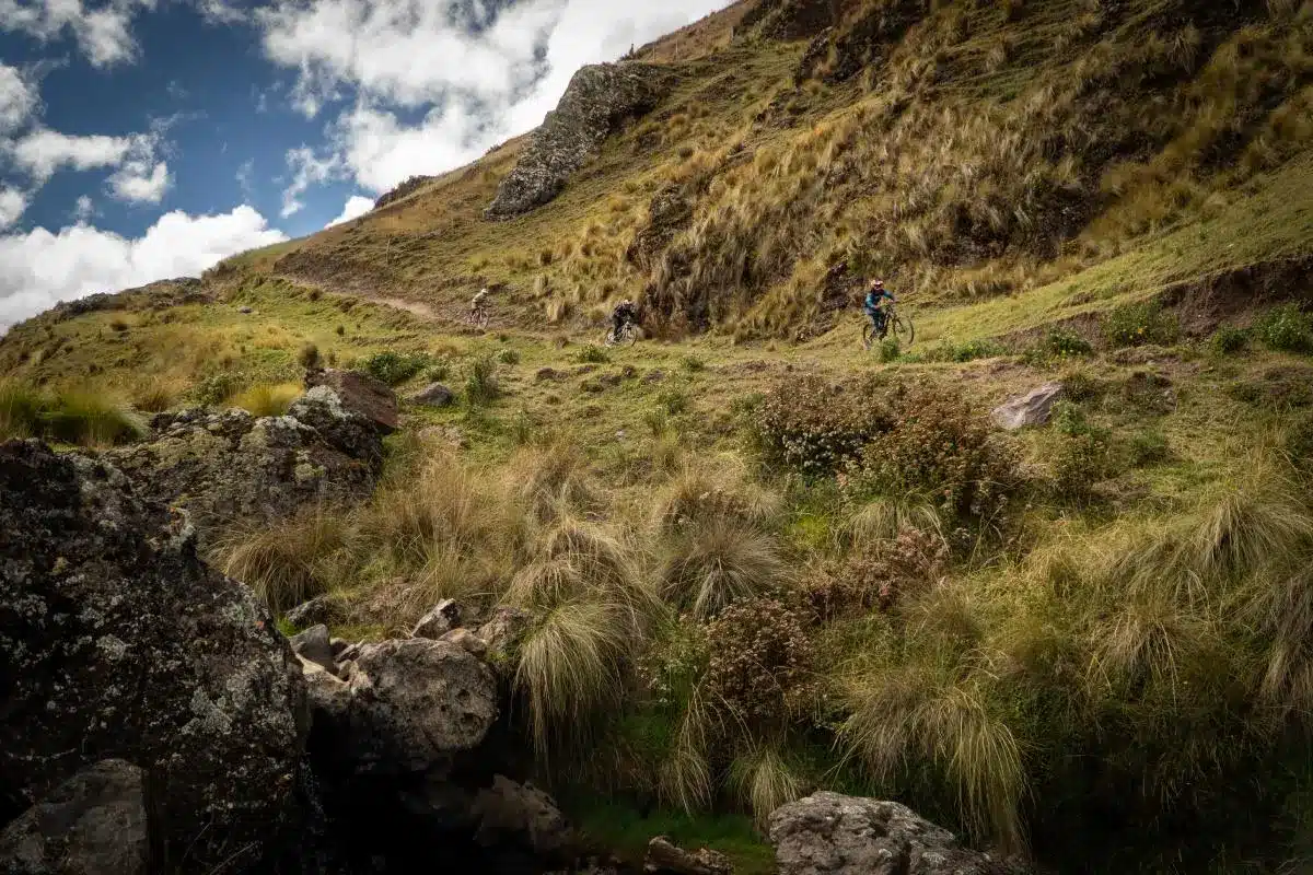 Ciclista recorriendo un estrecho sendero andino del circuito Lares con valles verdes, formaciones rocosas y nubes sobre las alturas de Cusco (Peru Biking Tours).