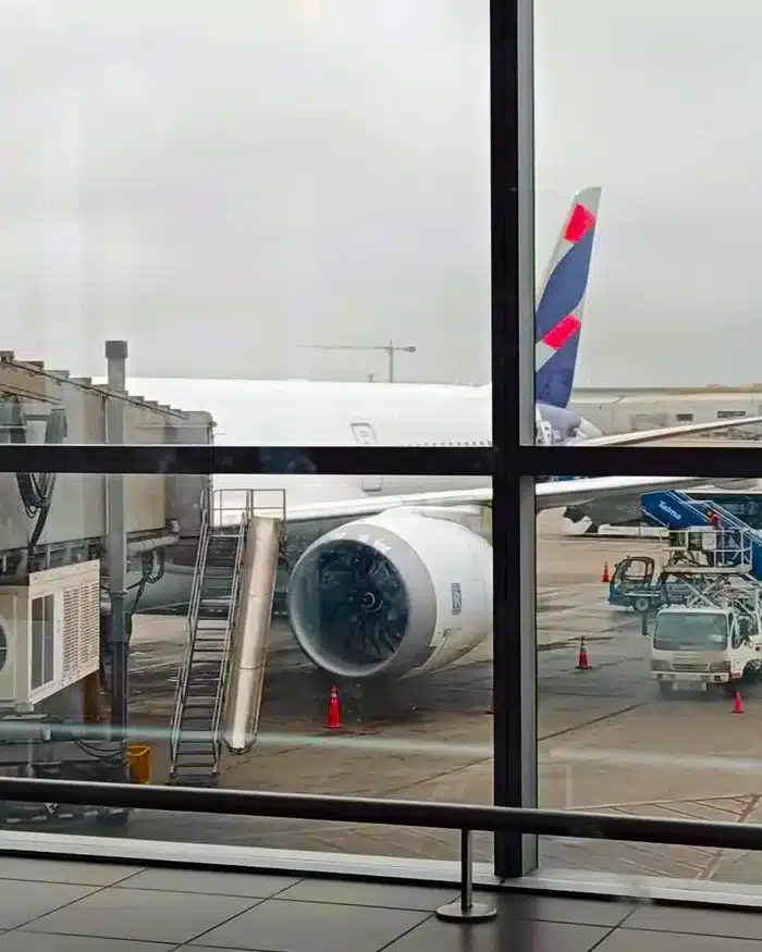 La imagen muestra un avión de la aerolínea LATAM Airlines estacionado junto a una manga de embarque en un aeropuerto internacional.