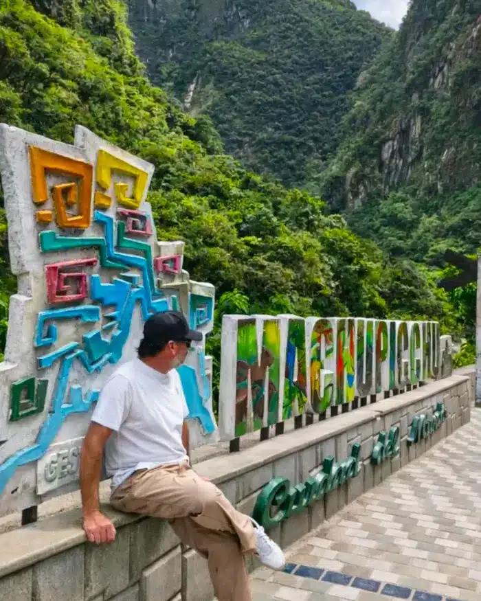 La imagen muestra a un viajero descansando junto a un letrero de Machu Picchu, rodeado de exuberante vegetación andina.