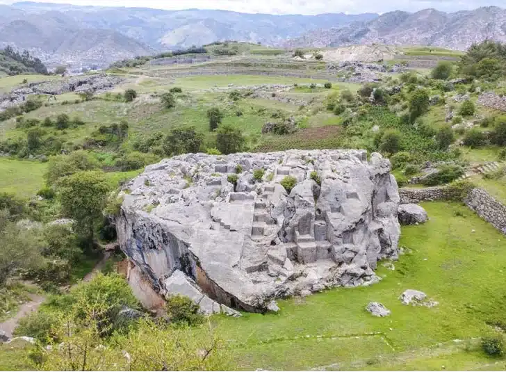View of the Great Chincana of Sacsayhuamán in Cusco, with carved rock formations, green terraces and Andean mountains.