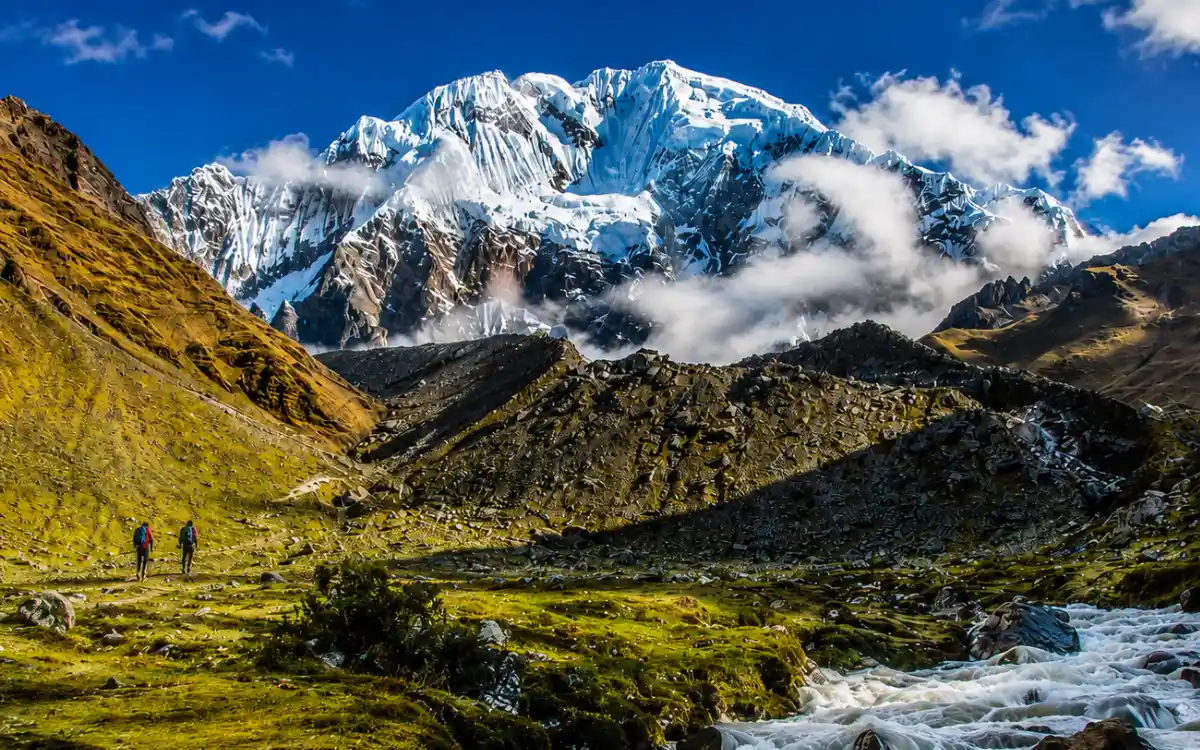 Imagen panorámica de la ruta hacia el apu Salkantay, río de montaña y dos viajeros.