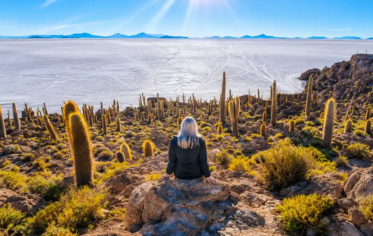 Mujer sentada entre cactus en la Isla Incahuasi, observando el Salar de Uyuni bajo un cielo despejado.