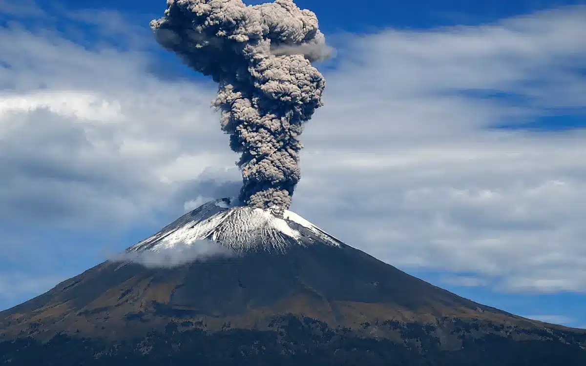 Volcán Popocatépetl con columna de humo y nieve en la cima, visto bajo cielo despejado con nubes.