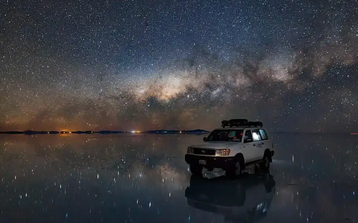 Auto todoterreno sobre el Salar de Uyuni con cielo estrellado, Vía Láctea y reflejo nocturno en el agua.