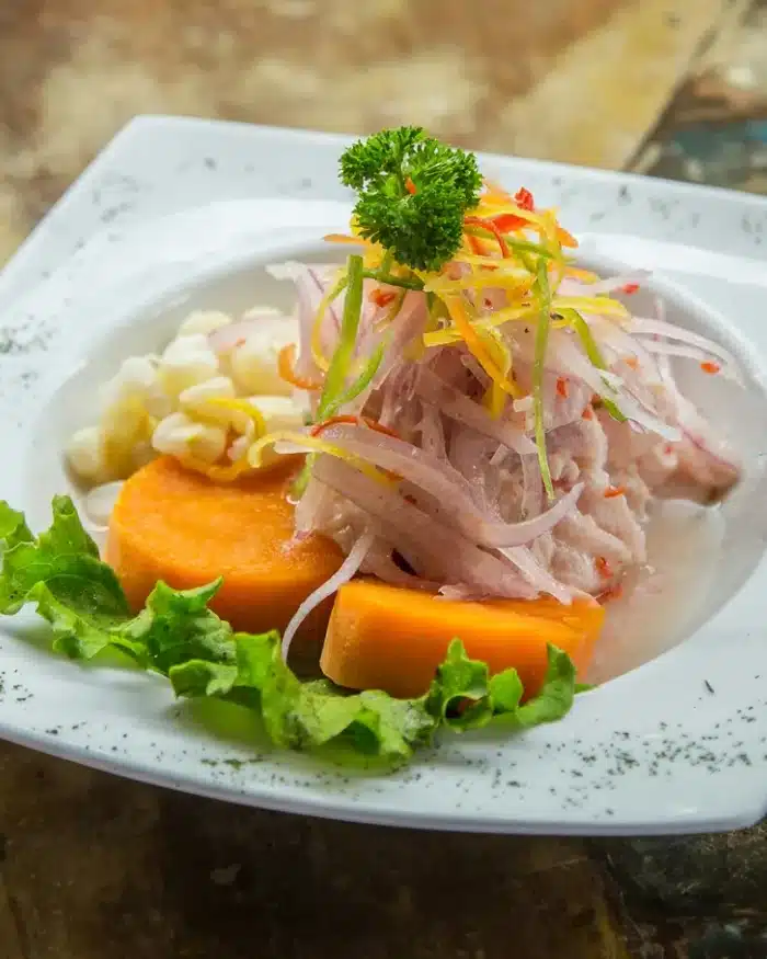 Ceviche peruano servido en plato blanco con camote, choclo, cebolla morada, lechuga y decoración de perejil.
