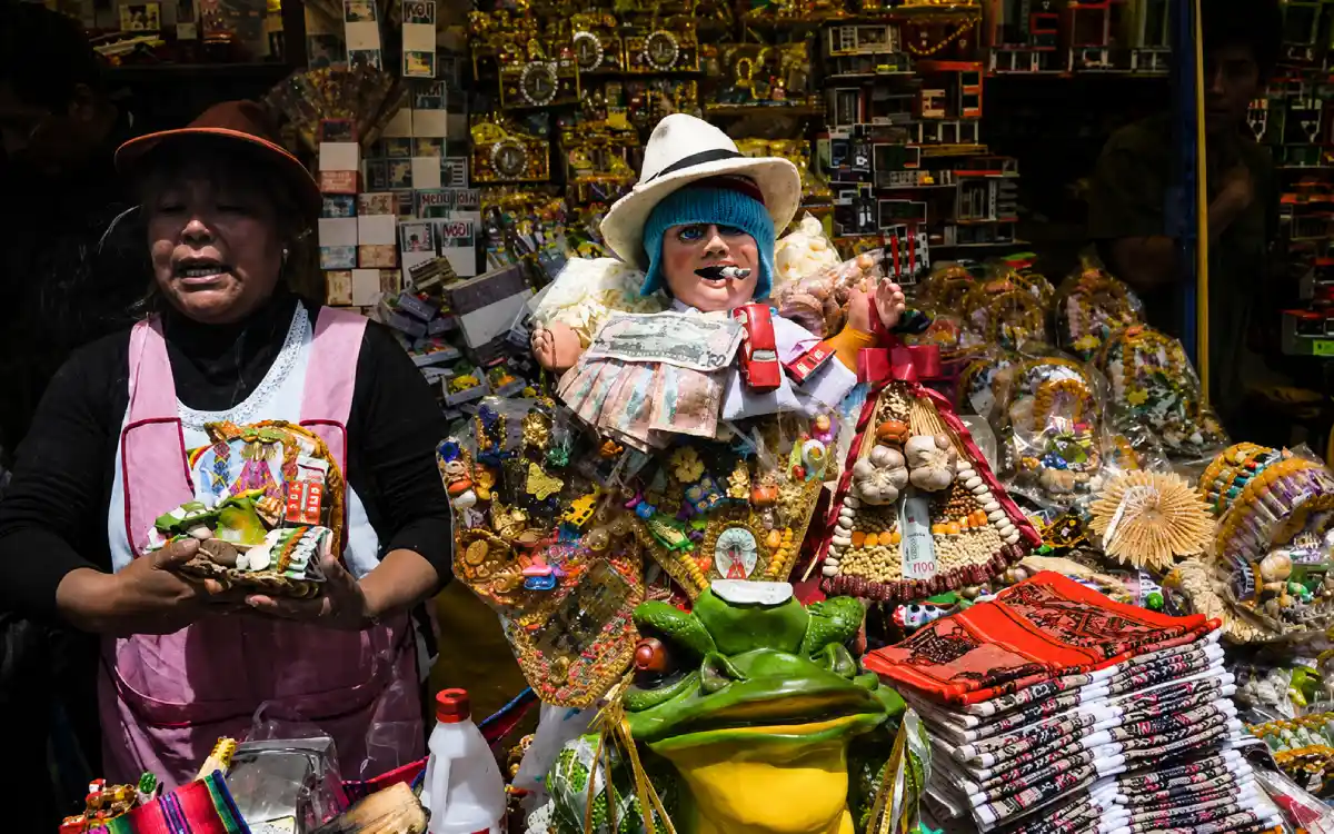 Figura del Ekeko en un puesto tradicional andino, rodeada de miniaturas, billetes, alimentos y objetos de abundancia.