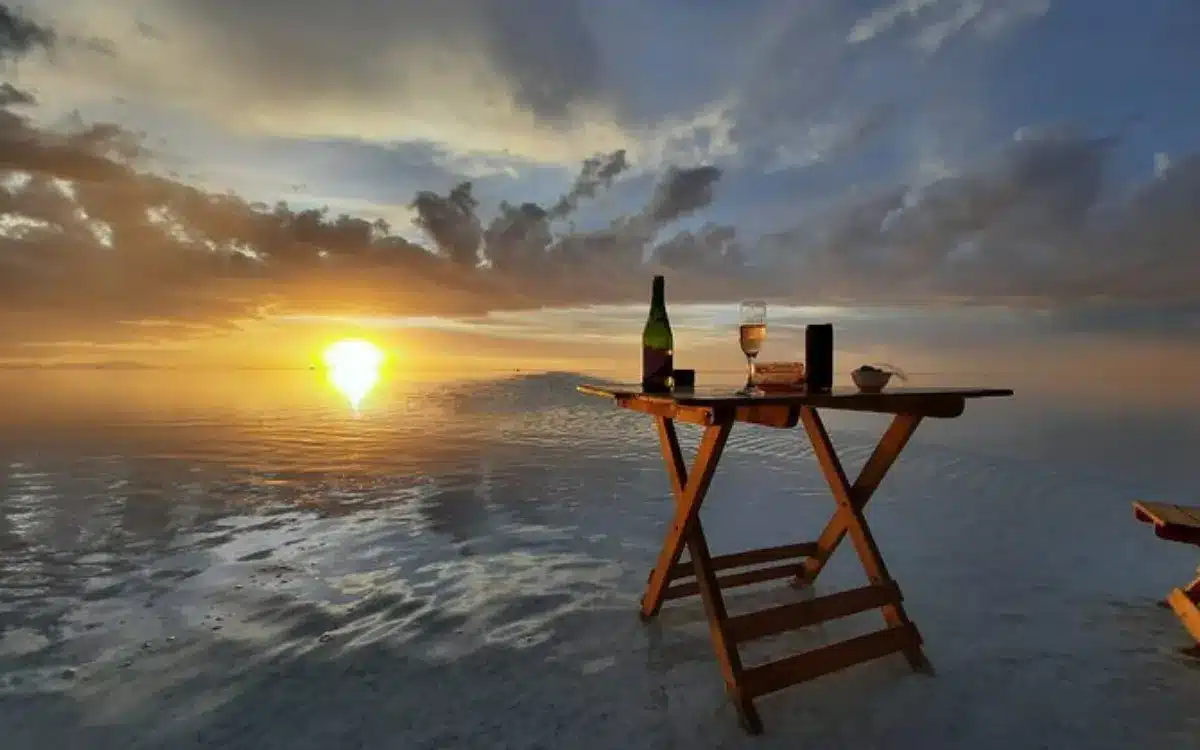 Mesa con vino y copas sobre el Salar de Uyuni durante el atardecer, con reflejo del sol en el agua.