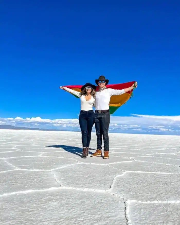 Pareja de viajeros sosteniendo bandera de Bolivia en el Salar de Uyuni, con cielo azul durante temporada seca.