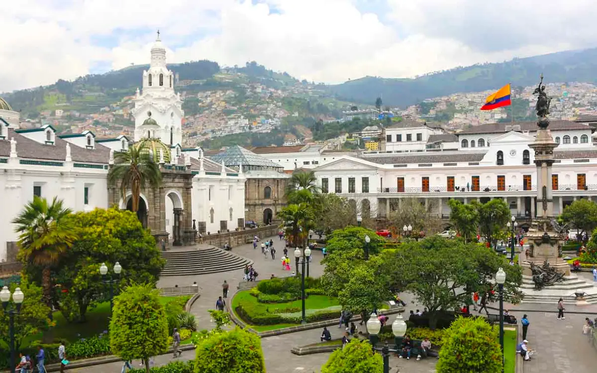 Plaza Mayor de Quito en Ecuador, con bandera nacional, estatua central, edificios coloniales y montañas al fondo.
