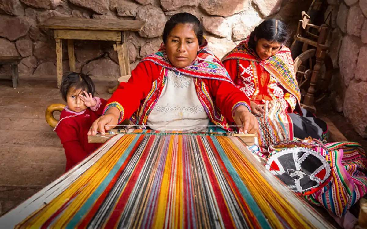 Tejedora artesanal en Awana Kancha realizando un tejido tradicional con lana de colores junto a mujeres con vestimenta andina.