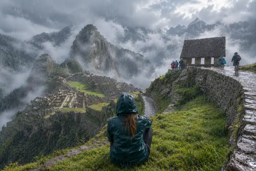 Traveler in a rain jacket overlooks Machu Picchu during heavy rain, with wet paths, Inca ruins and mountains partly covered by mist.