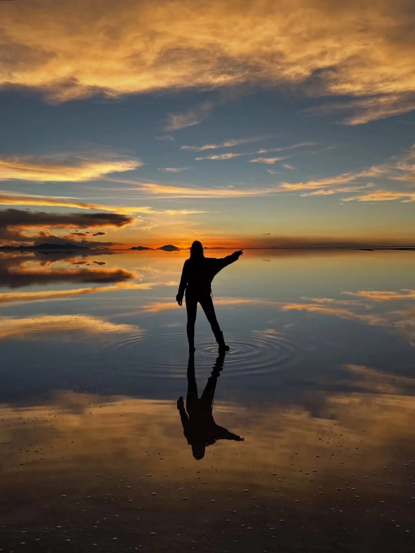 Traveler watches sunrise over the flooded Uyuni Salt Flat, with the sky and her silhouette reflected across the mirror-like surface.
